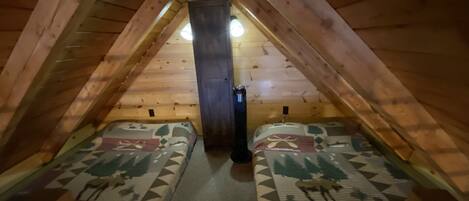 A small, cozy attic bedroom with wooden walls and a sloped ceiling. There are two beds with patterned bedding and a small window.