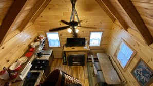 Une cabane confortable avec un intérieur en bois, un ventilateur de plafond, un coin cuisine équipé d’une cuisinière et d’un évier, et deux fenêtres avec des rideaux.