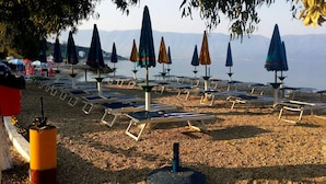 A beach with rows of sun loungers and umbrellas, a rocky shoreline, and mountains in the background. - Villaggio shehu (Orikum)