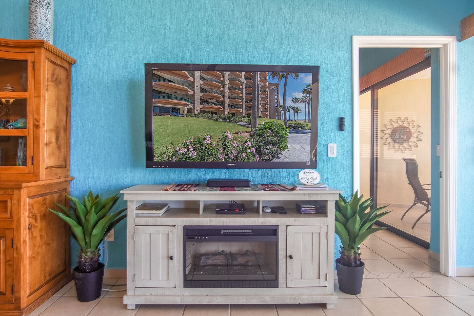 A living room with a flat-screen TV mounted on a wall, a wooden cabinet with glass doors, a fireplace, and two potted plants.