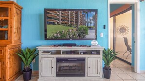 Una sala de estar con un televisor de pantalla plana montado en la pared, un mueble de madera con puertas de cristal, una chimenea y dos plantas en macetas.