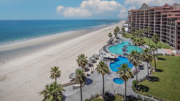 Un complejo turístico con una amplia zona de piscina, una playa y un edificio de hotel con varios balcones.