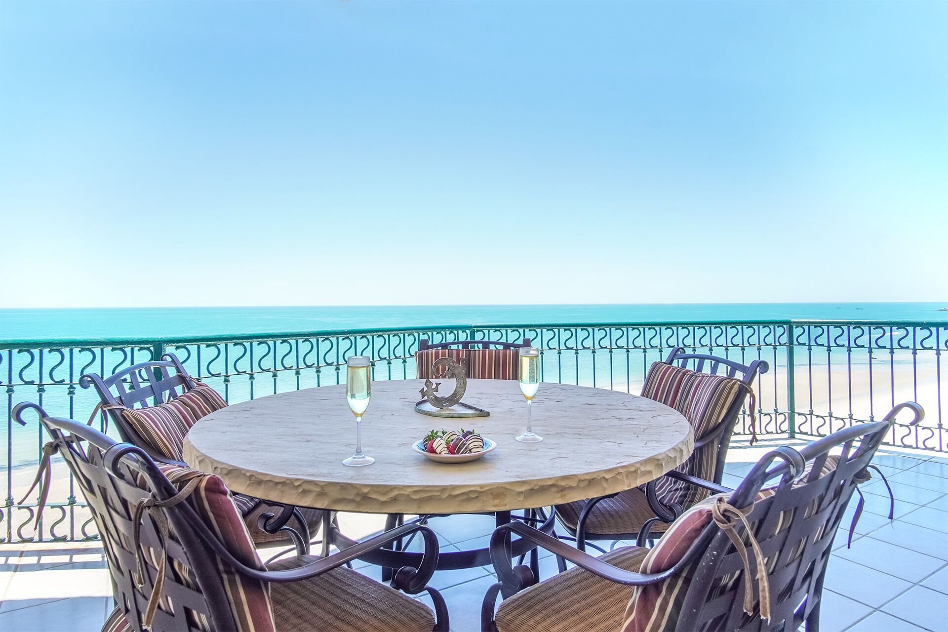 A round table with six wicker chairs, two wine glasses, and a plate of strawberries on a balcony overlooking the sea.