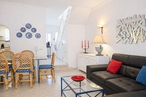 A living room with a sofa, chairs, a glass coffee table, a dining area with a table and chairs, a staircase, and wall decor.