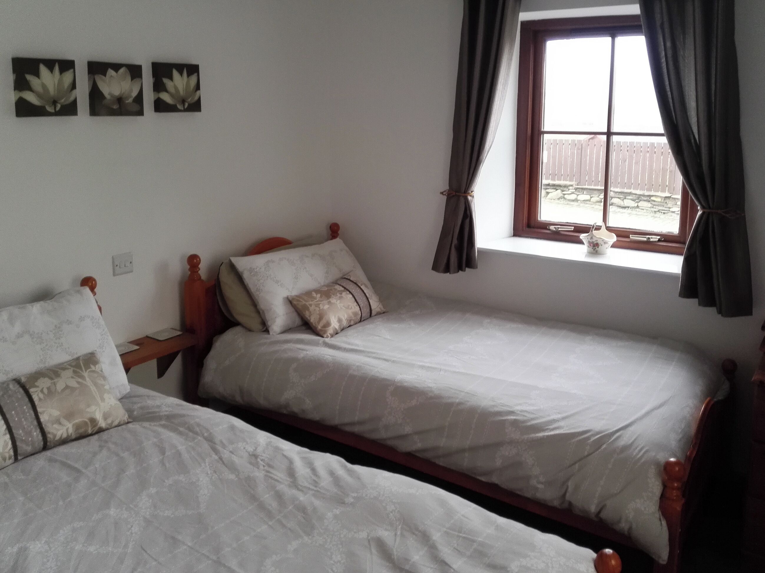 Une chambre avec deux lits simples, une fenêtre avec des rideaux et trois tableaux représentant des fleurs, accrochés au mur.