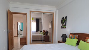 A bedroom with a large bed, a wardrobe with a mirror, a bedside table with a lamp, and a wall with a single piece of artwork. - T3, espaçoso, wifi, a/c, junto à praia! (Albufeira)
