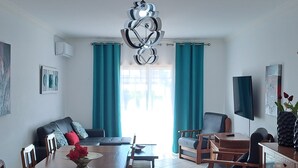 A modern living room with a dining area, teal curtains, a black sofa, a wooden dining table, and a TV on the wall. - T3, espaçoso, wifi, a/c, junto à praia! (Albufeira)