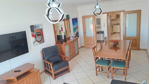 A modern living room with a TV, wooden furniture, a dining table with chairs, and two pendant lights. - T3, espaçoso, wifi, a/c, junto à praia! (Albufeira)