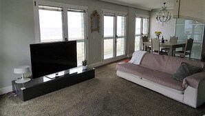 A living room with a large flat-screen TV on a stand, a white sofa, a dining area with a table and chairs, and large windows offering a view. - Duplex San Luis - 1ªline beach, incredible view garage by Egona (Zarautz)