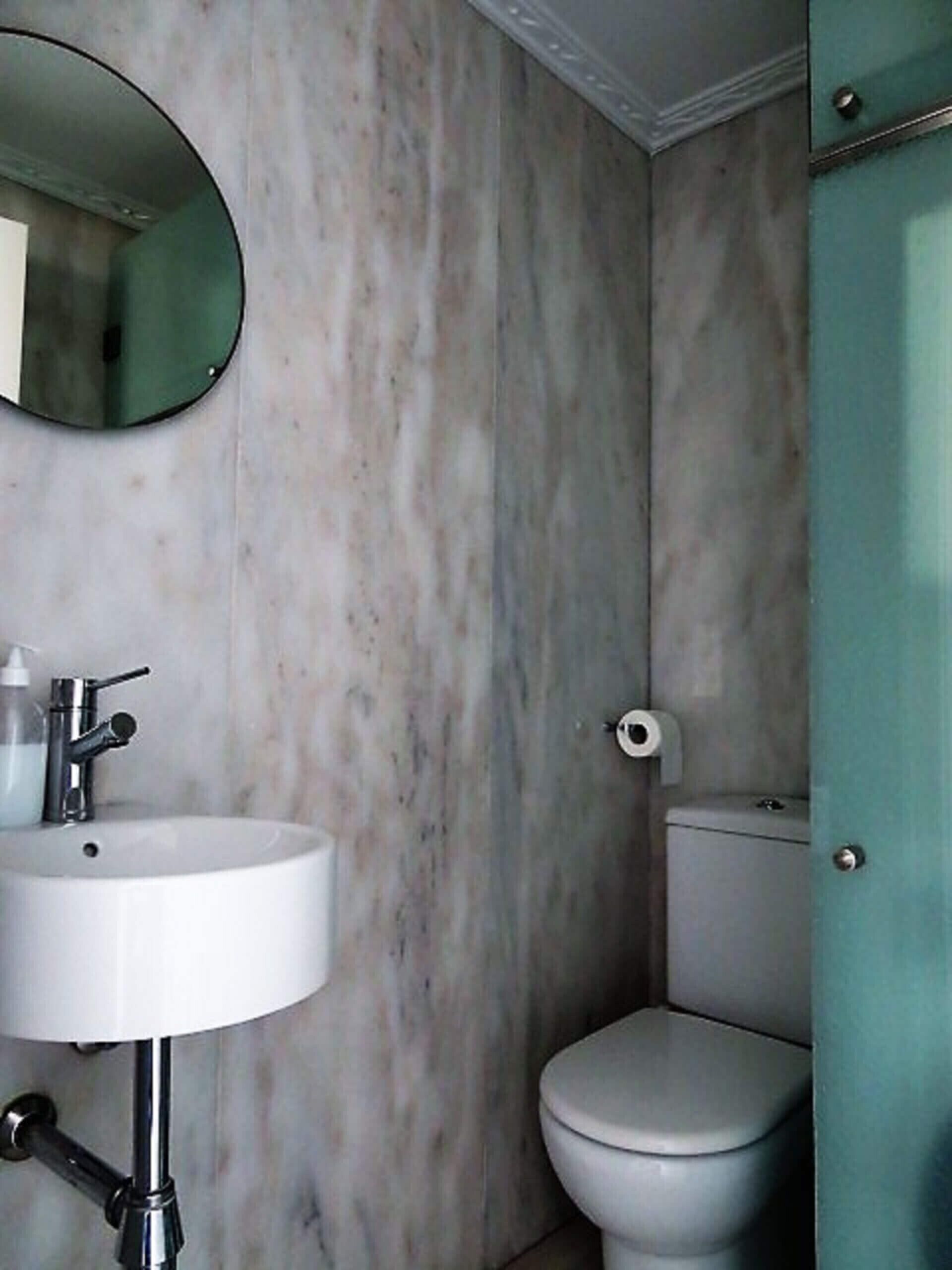 Un baño con pared de mármol, un lavamanos blanco con grifo cromado, un espejo redondo y un inodoro.