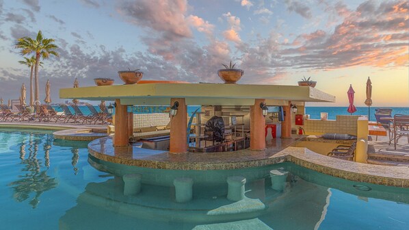Ein Poolbereich mit Liegestühlen, einer überdachten Sitzgelegenheit und Blick auf das Meer bei Sonnenuntergang.