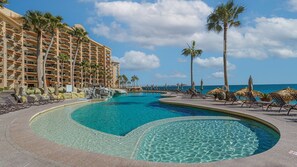 A resort pool with clear blue water, surrounded by lounge chairs and palm trees, with a hotel building in the background.