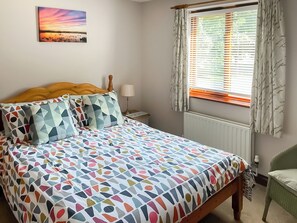 A bedroom with a bed, a nightstand, a lamp, a window with curtains, and a wall-mounted picture.