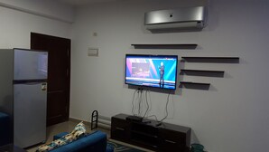 Una sala moderna con un televisor de pantalla plana montado en la pared, aire acondicionado integrado, un refrigerador y un mueble de entretenimiento de madera oscura.