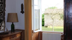 A room with a wooden dresser, a lamp, a window with curtains, and a view of a garden and stone wall. - Logis near the chateau (Paizay-Naudouin-Embourie)