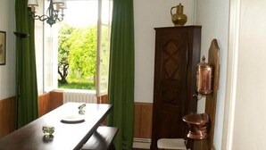 A dining area with a wooden table, chairs, a green window with curtains, a chandelier, and a cabinet with a copper pot. - Logis near the chateau (Paizay-Naudouin-Embourie)