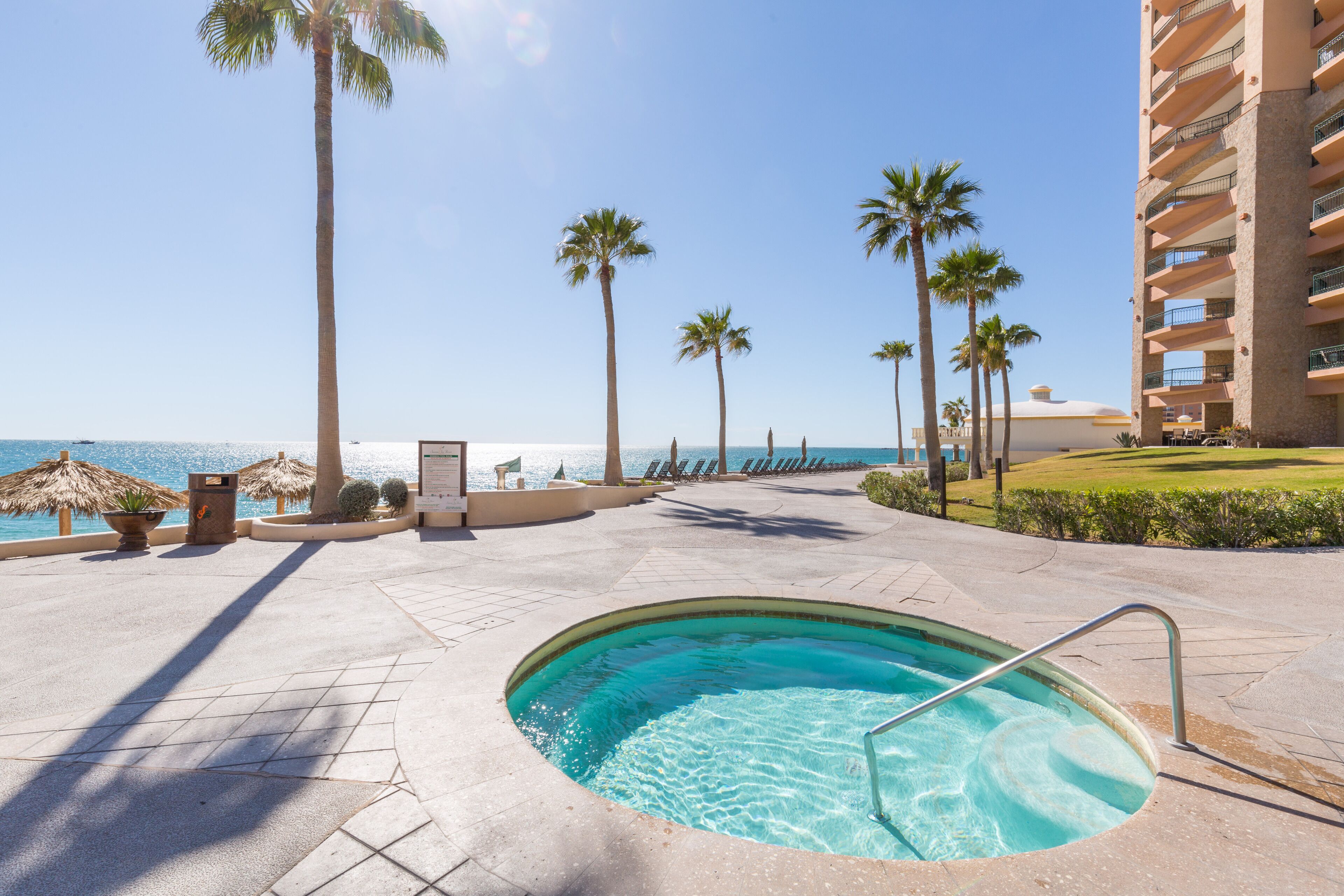 A pool area with a clear view of the ocean, palm trees, and a hotel building.