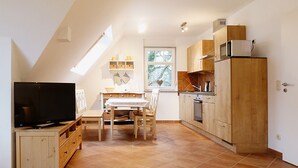 A compact kitchen with a dining area, a TV, and a window with a view of trees. - cozy apartment near the Darßwald, beach chair on balcony, WiFi, 4 pers. (Prerow)