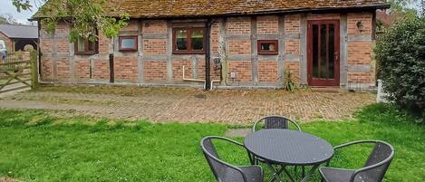 A brick building with a tiled roof, a red door, and a small window. There is a paved area with a table and chairs in front.