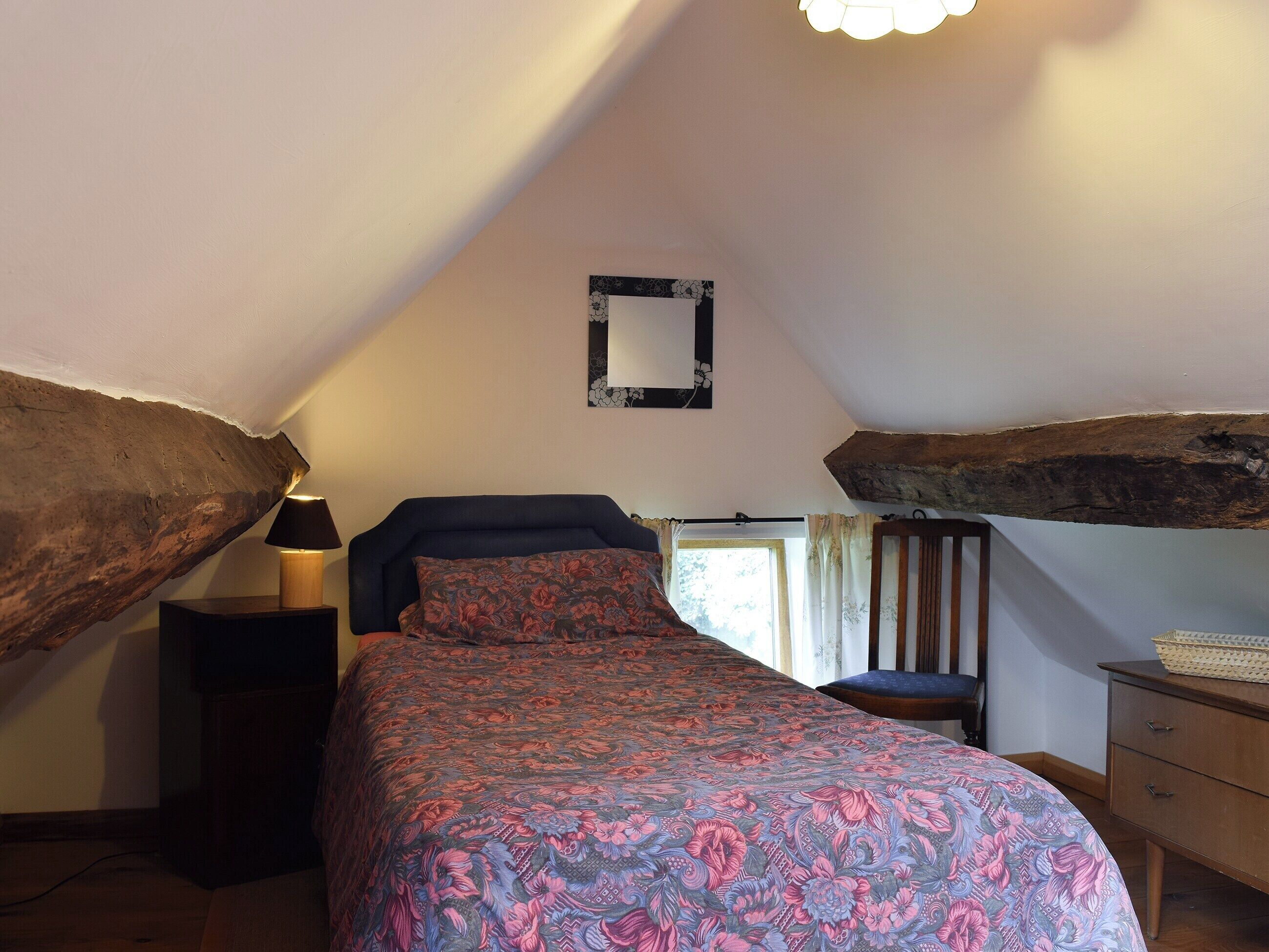 Une chambre avec un plafond en pente, un lit recouvert d’une couvre-lit à motifs floraux, une table de chevet, une chaise et une commode.