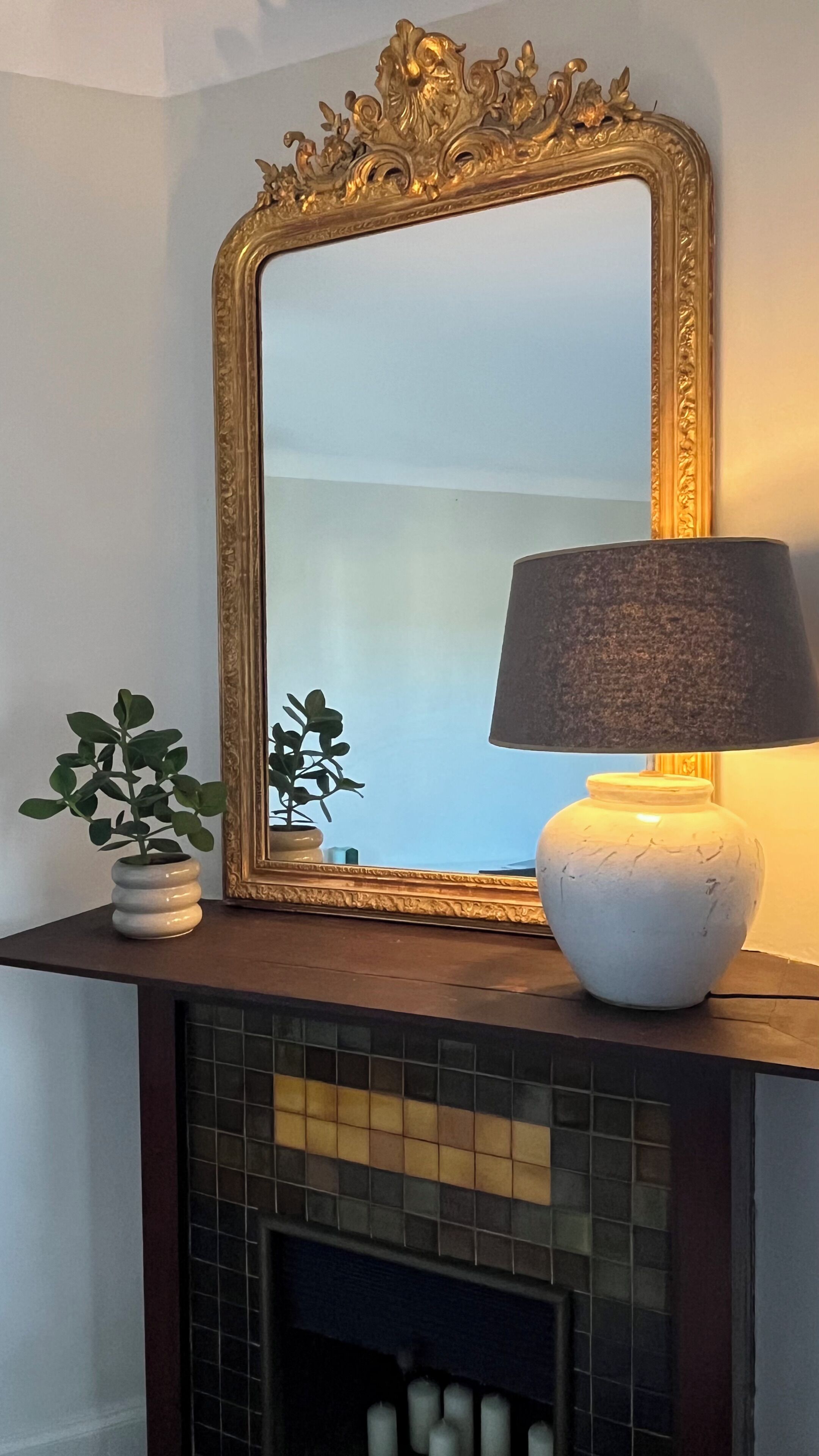 A mirror with an ornate gold frame, a white lamp with a dark shade, and a small potted plant on a mantelpiece above a tiled fireplace.