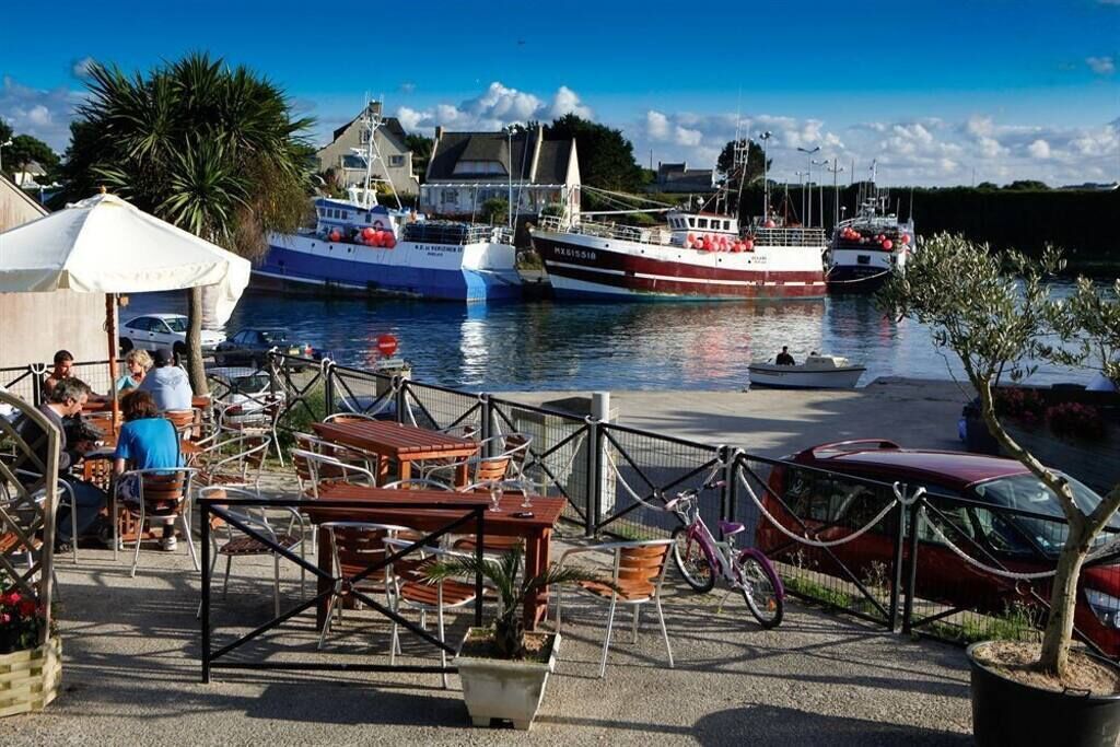 A waterfront dining area with tables and chairs, boats docked in the background, and a red car parked nearby.