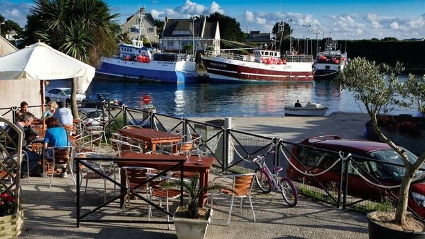 A waterfront dining area with tables and chairs, boats docked in the background, and a red car parked nearby.