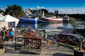 Ein Speisebereich am Wasser mit Tischen und Stühlen, im Hintergrund liegen Boote im Hafen und in der Nähe ist ein rotes Auto geparkt.