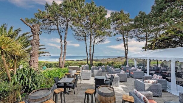 Ein Außenbereich mit Korbmöbeln, Holztischen und Fässern mit Blick auf Strand und Meer.