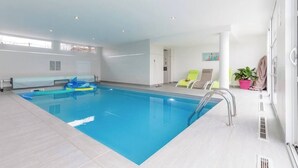 Una piscina interior con agua azul cristalina, un trampolín y un área de descanso con sillones y una planta en maceta.