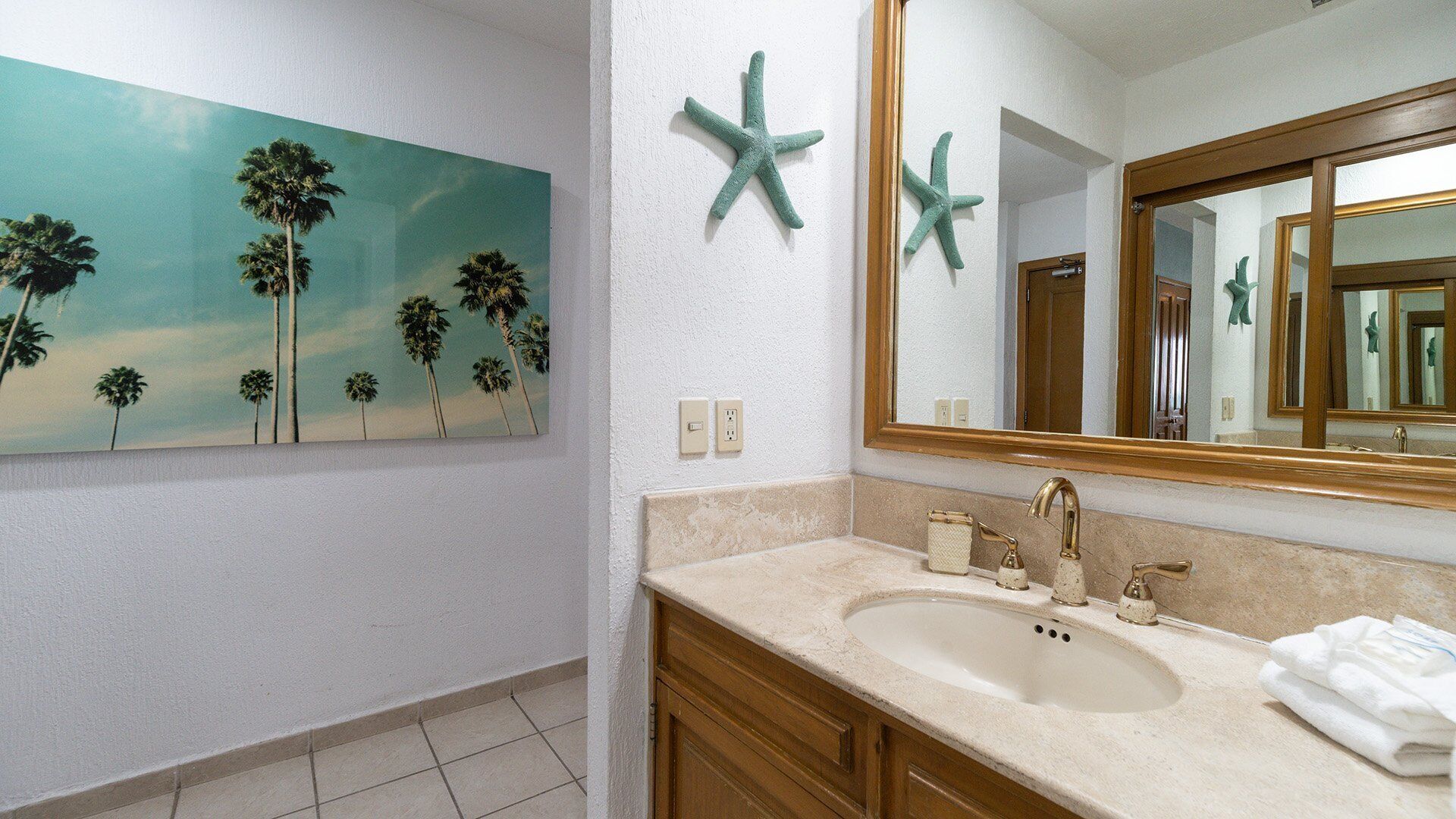 A bathroom with a large painting of palm trees, a sink with a gold faucet, and two starfish decorations on the wall.