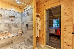 Bagno di una baita in legno con doccia, una panca con asciugamani e un letto visibile attraverso una porta aperta.