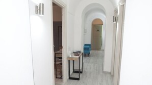 A hallway with white walls, a wooden floor, and a blue chair. There is a table with a lamp and a book. - Cineroma Guesthouse (Rome)