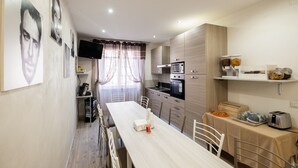 A modern kitchen with a dining area, featuring a long table, chairs, a microwave, oven, and a coffee machine. - Cineroma Guesthouse (Rome)