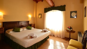 Une chambre d’hôtel avec un grand lit, une tête de lit en bois, des rideaux verts, une petite table ronde ornée d’un vase de fleurs et une chaise.