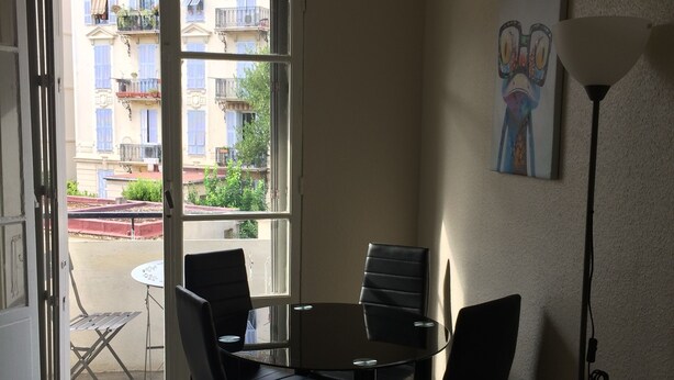 A room with a round table and four chairs, a water bottle on the floor, a painting on the wall, and a view of buildings through an open door. - Charming two room city center (Nice)