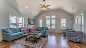 A spacious living room with a high ceiling, a ceiling fan, and large windows offering a view of the outside. - 4746- Sandy Feet Retreat - Oceanside Kitty Hawk Retreat w/ Pool (Kitty Hawk)