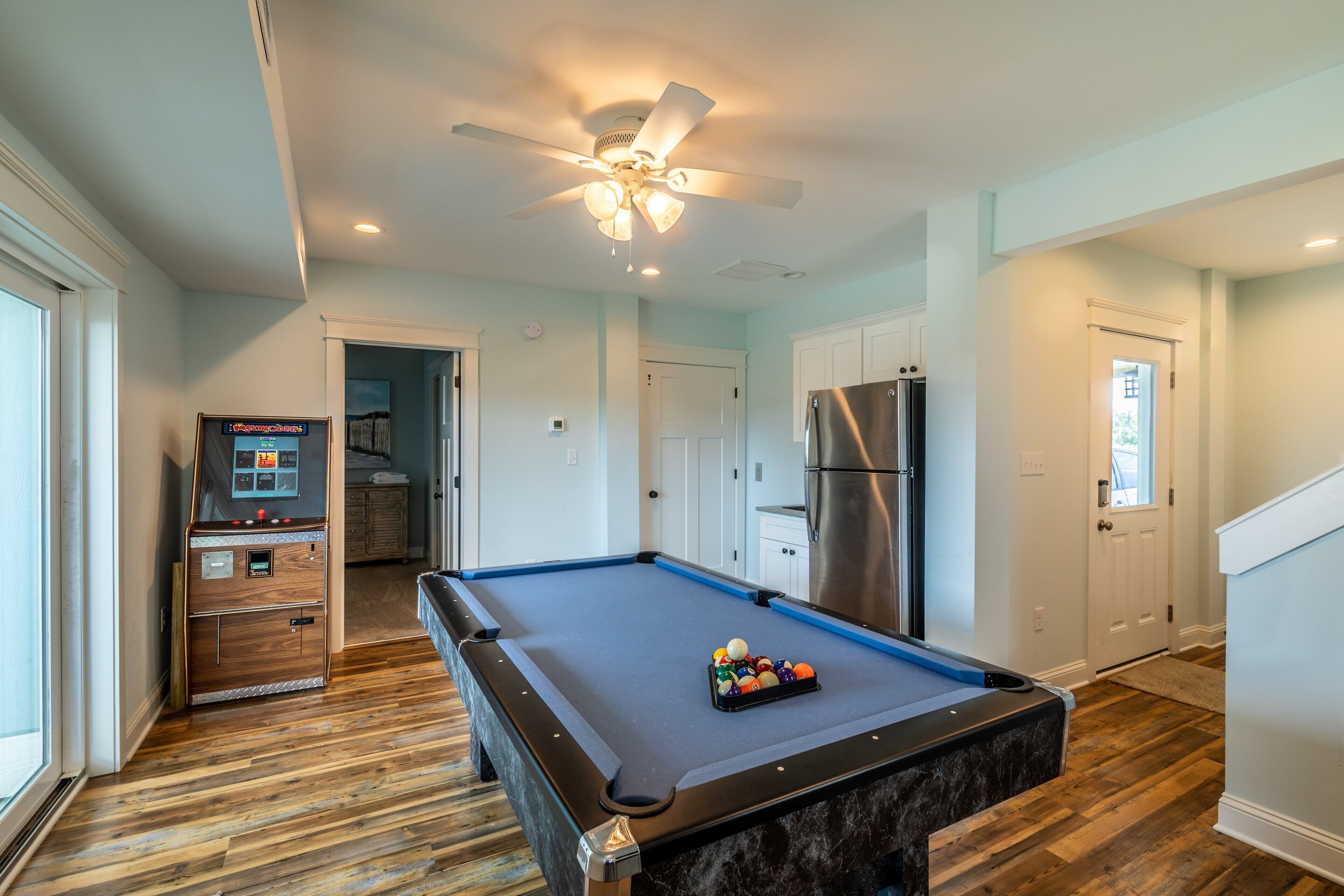 A pool table with balls, a ceiling fan, and a refrigerator in a room with wooden flooring and white walls.