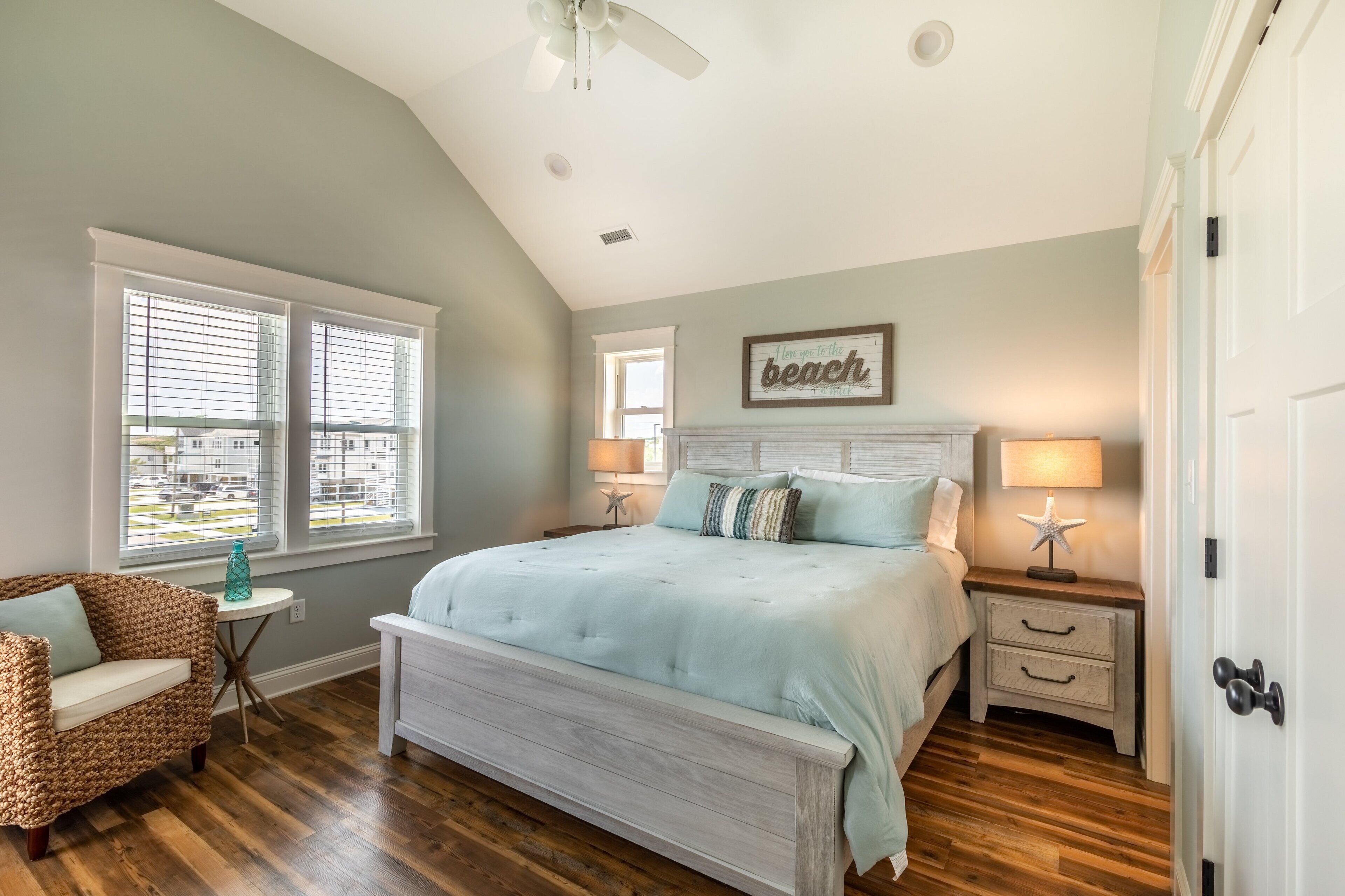 A bedroom with a large bed, two bedside tables with lamps, a wicker chair, and a window with blinds.