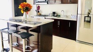 Cozinha moderna com armários de madeira escura, geladeira de aço inoxidável, micro-ondas e fogão a gás. Há uma pequena ilha com banquetas e um vaso de flores.