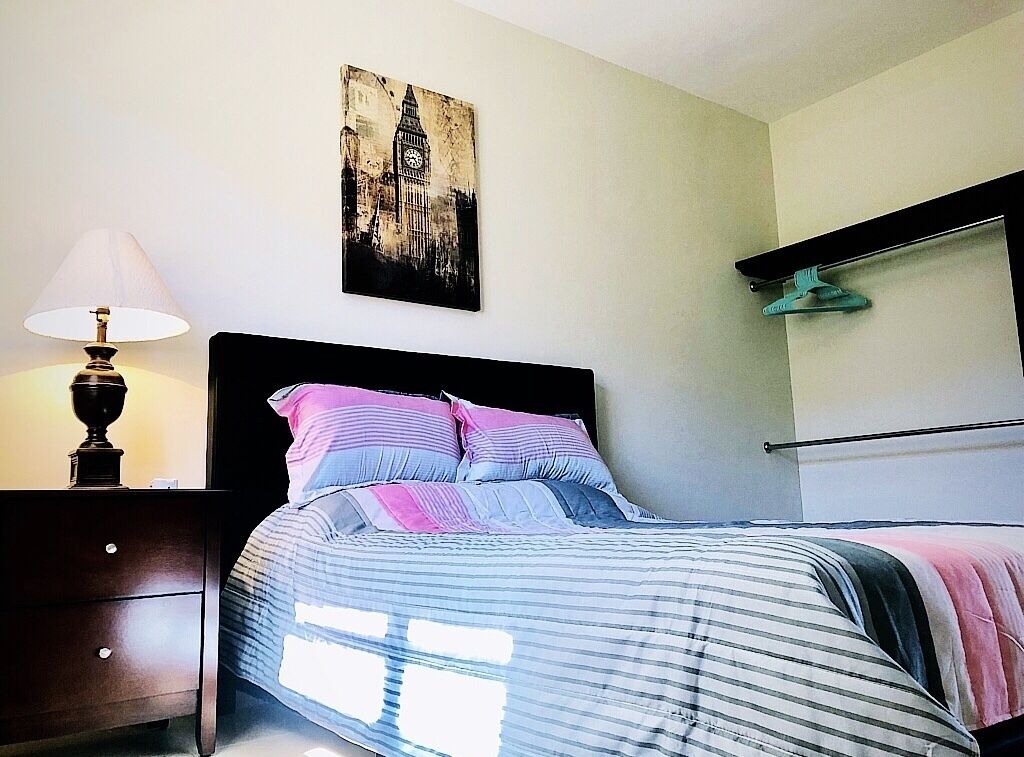 A bedroom with a bed, a nightstand with a lamp, a wall-mounted shelf, and a picture of a building.