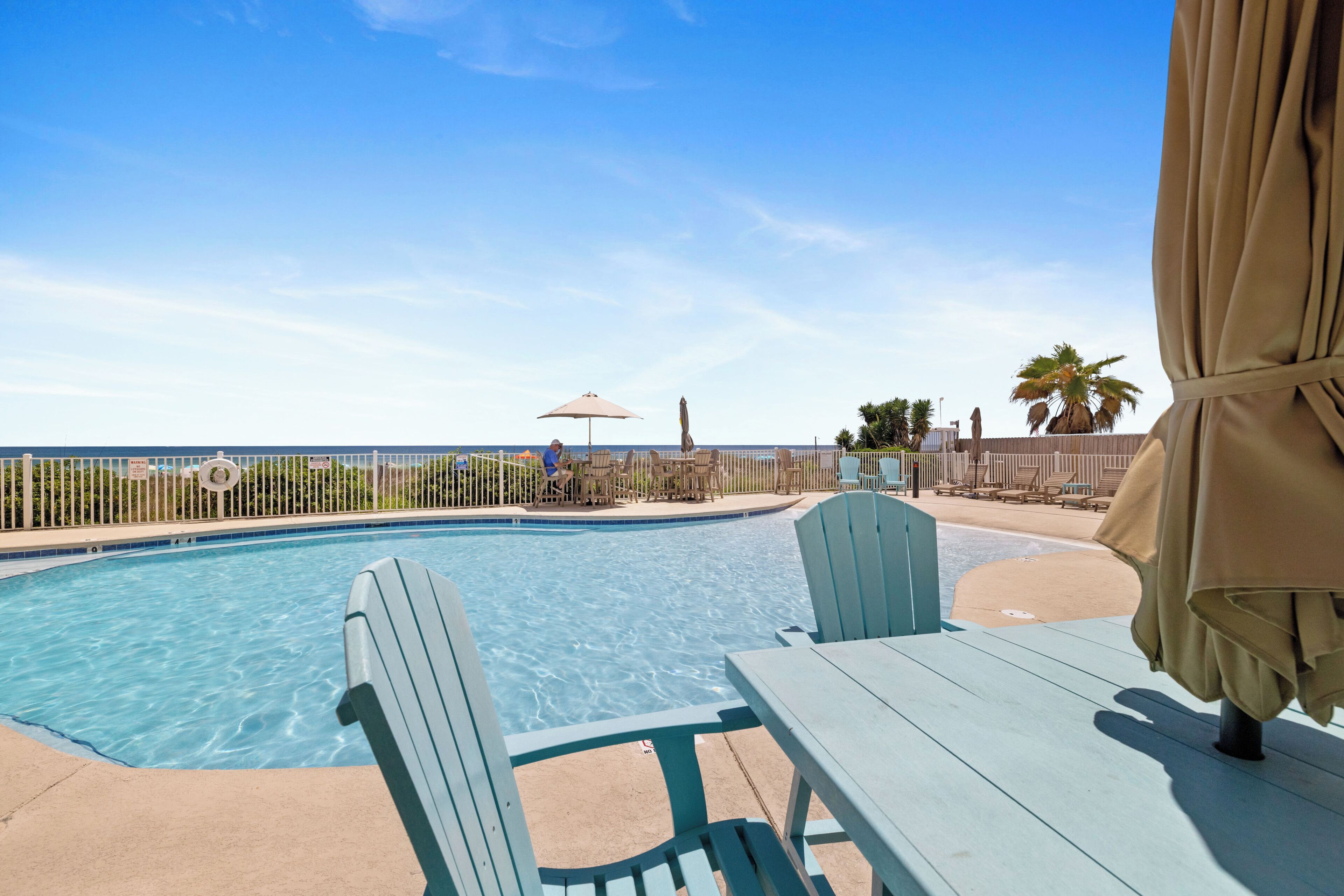 A swimming pool with a view of the beach, surrounded by a fence, and a table with a chair.
