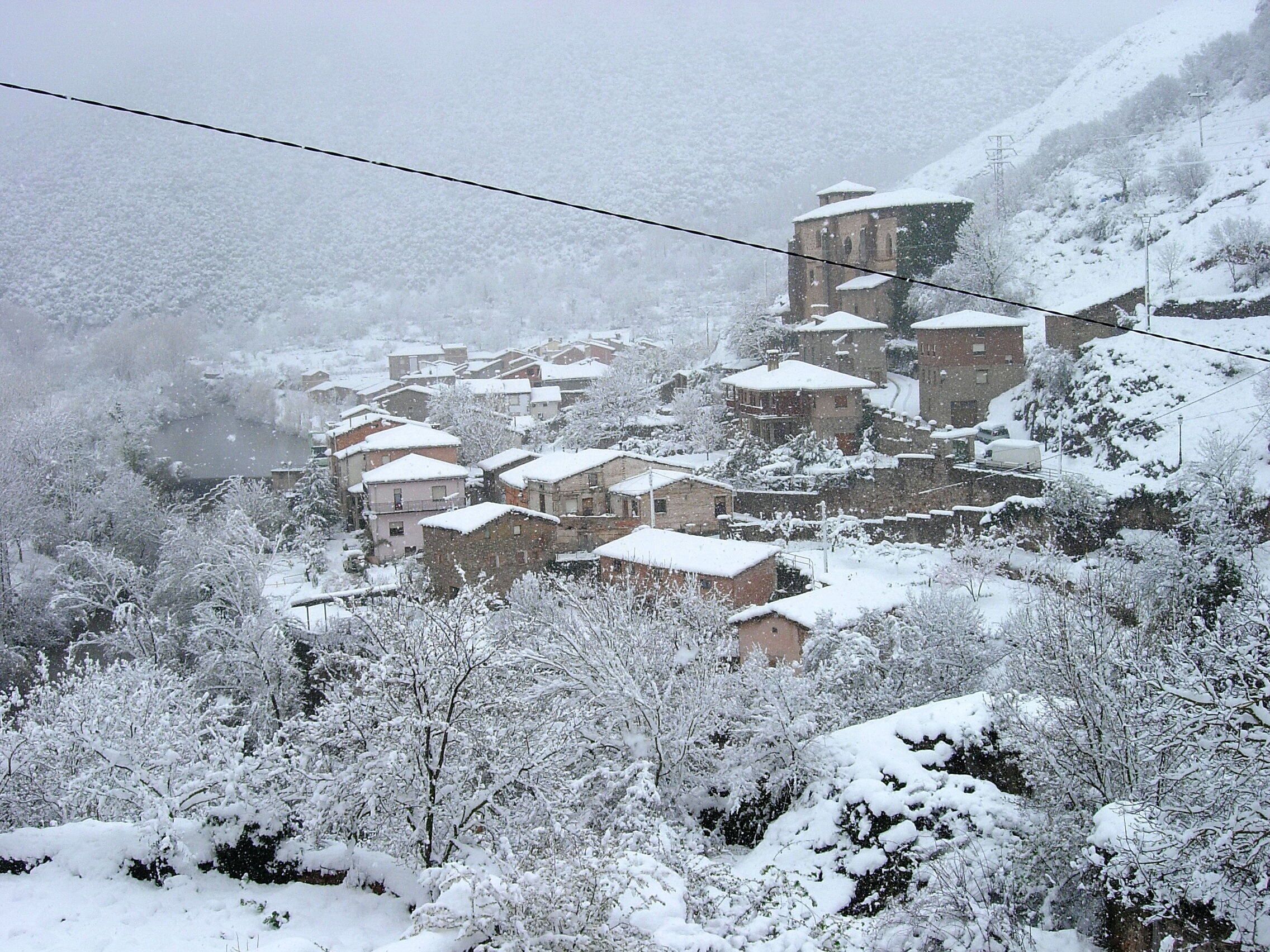 A snow-covered village with traditional stone buildings, a church, and a hill in the background.