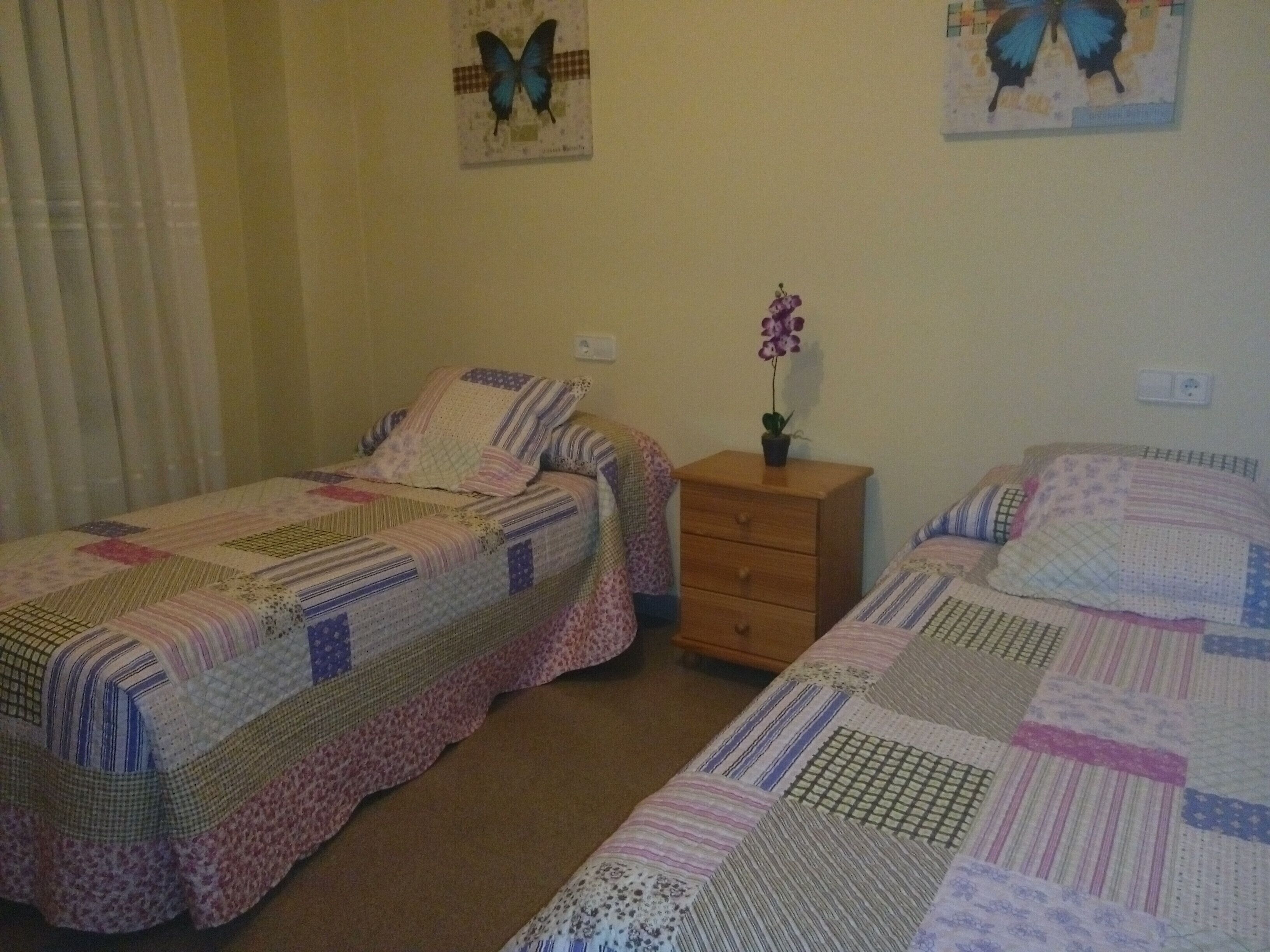 Un dormitorio con dos camas individuales, cada una con una colcha acolchada, una mesita de noche de madera con una orquídea en maceta e interruptor de luz en la pared.