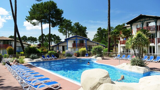 A swimming pool with lounge chairs, a building with balconies, and surrounding trees.