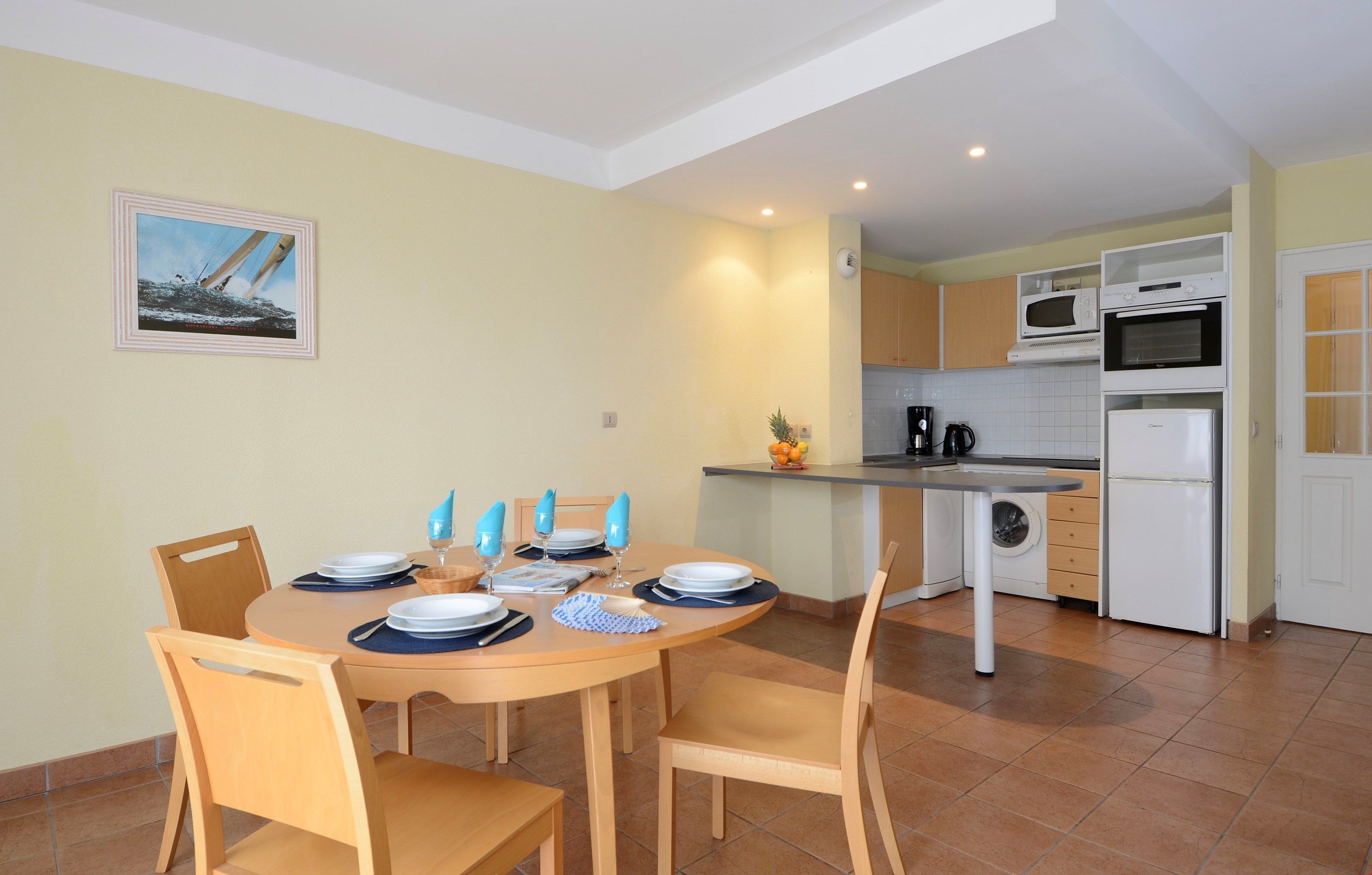 A dining area with a round table set for four, surrounded by wooden chairs. A kitchen with white appliances and a tiled backsplash is visible in the background.