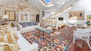 Un loft espacioso con pared de piedra, un sofá blanco, un tapete estampado y una zona de comedor con sillas de mimbre.