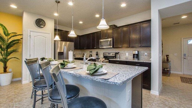 A kitchen with a central island, dark cabinets, stainless steel appliances, and pendant lighting. - 5BR Windsor Townhome | Private Splash Pool | Sleeps 12 | Dog Friendly | Near Disney (kissimmee)