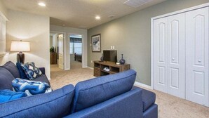 A living room with a sofa, a television, and a cabinet. There is a hallway leading to a door. - 5BR Windsor Townhome | Private Splash Pool | Sleeps 12 | Dog Friendly | Near Disney (kissimmee)