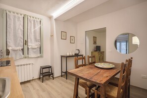 A dining area with a wooden table, chairs, a small stool, a radiator, a window with curtains, a table with teapots, a mirror, and framed pictures on the wall.
